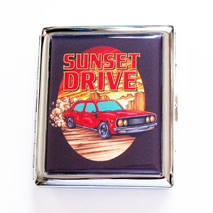 Pode incluir: Um porta-cigarros retangular de metal prateado com um gráfico de um carro vermelho numa estrada deserta e as palavras "SUNSET DRIVE" em laranja. O fundo é preto e o porta-cigarros tem um acabamento brilhante.