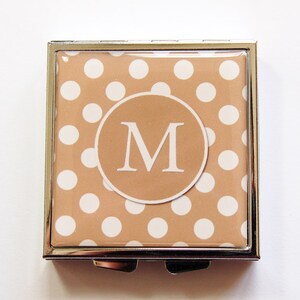 Può includere: Un specchietto compatto quadrato con cornice argentata. La parte superiore presenta uno sfondo beige a pois bianchi e un design circolare con la lettera "M". Il compatto è chiuso.