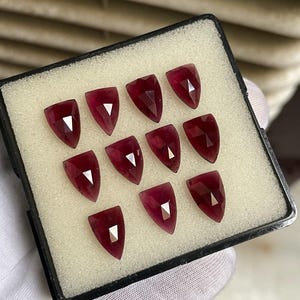 Könnte beinhalten: Eine Sammlung von zehn tiefroten, schildförmigen facettierten Edelsteinen, die in einem Raster in einer schwarzen Präsentationsbox mit weißer Schaumstoffeinlage angeordnet sind. Jeder Stein hat eine Spitze und mehrere Facetten.