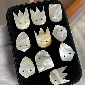 May include: A collection of ten iridescent mother-of-pearl buttons arranged on a black rectangular display card. The buttons feature various shapes, including crown-like points and rounded edges, each with two small holes for sewing attachment.