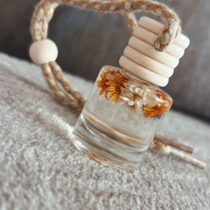 May include: A small, clear glass bottle filled with a clear liquid and dried orange flowers. The bottle has a wooden cap and is suspended by a beige and gold rope. The bottle is designed to be hung, likely as an air freshener.
