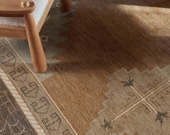 May include: A close-up of a brown and beige patterned rug with a geometric design. The rug has a border with a repeating pattern and is partially covered by a wooden chair with a white seat.