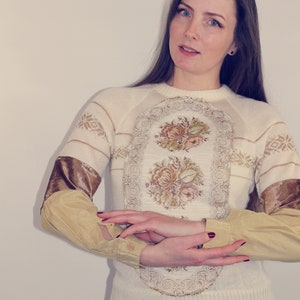 Könnte beinhalten: Cremefarbener Pullover mit floralem Design und braunen Samtakzenten an den Ärmeln. Der Pullover hat einen runden Ausschnitt und wird mit einem hellgelben Button-Down-Hemd darunter kombiniert. Das Logo The Bohemian Dream befindet sich in der oberen linken Ecke.