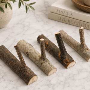 May include: A set of five rustic wooden coat hooks. Each hook is made from a section of a tree branch, with a smaller branch extending upwards to serve as the hook. The wood tones vary from light tan to dark brown.