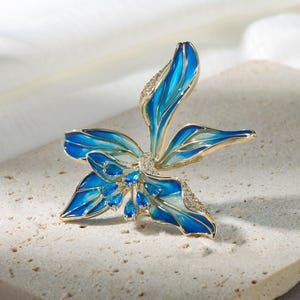 May include: A gold-toned floral brooch with blue enamel petals and clear crystal accents. The brooch has a detailed design with multiple layers of petals and small blue crystal embellishments. The brooch is set on a textured surface.