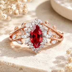 Op de afbeelding: Een roségouden ring met een centrale marquise-geslepen rode edelsteen, omringd door een halo van kleine witte steentjes. De ringband is voorzien van verfijnde metalen bladmotieven die uitwaaieren vanaf de centrale zetting.