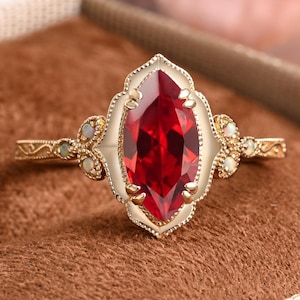 May include: A gold ring featuring a large marquise-cut deep red gemstone at the center, surrounded by a scalloped metal border. The band is adorned with small, round, iridescent white opals on each side and intricate engraved scrollwork details.