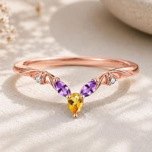 Puede incluir: Un anillo en forma de V de oro rosa con un citrino amarillo central en talla pera, dos amatistas moradas en talla marquesa y dos pequeños diamantes blancos redondos engastados en detalles metálicos en forma de ala a cada lado.
