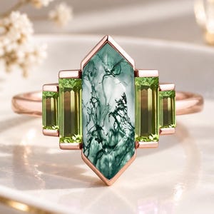 May include: A rose gold ring featuring a central elongated hexagonal moss agate stone with dark green dendritic inclusions, flanked by two pairs of vertical baguette-cut light green peridot stones set in a symmetrical geometric arrangement.