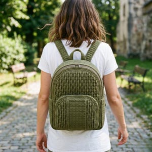 May include: Olive green backpack with a woven pattern, featuring a zippered main compartment and a front pocket. The backpack has adjustable straps and a top handle. The bag is made of a textured material.