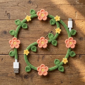 May include: A crocheted USB charging cable decorated with green vines, leaves, and peach and yellow flowers. The cable has a USB-A connector on one end and a USB-C connector on the other. The cable is arranged in a decorative floral pattern.