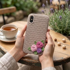 Könnte beinhalten: Eine beige gehäkelte Handyhülle mit floralem Design. Die Hülle hat ein strukturiertes Muster und ist mit drei Häkelblumen in Rosa- und Lilatönen sowie grünen Blättern verziert. Die Handyhülle wird von einer Person in den Händen gehalten.