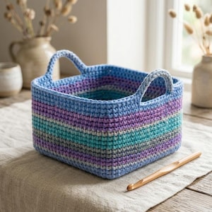 May include: A handmade, square crochet basket with two handles. The basket features horizontal stripes of blue, purple, teal, and gray. A wooden crochet hook rests on the neutral-colored fabric next to the basket.
