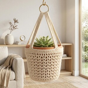 May include: A cream-colored crocheted plant hanger featuring an open-weave pattern and two sturdy straps that connect to a gold-toned metal ring. The hanger holds a terracotta pot containing a green succulent plant, suspended from a hook.