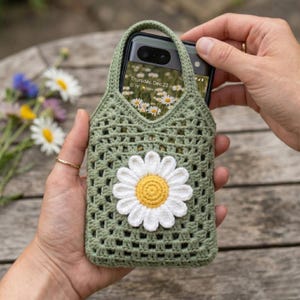 May include: A green crochet phone case featuring a white and yellow daisy. The case has a handle and a grid design. The phone screen shows a daisy field. The case is held by a person.