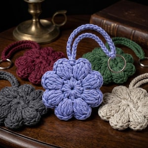 May include: A collection of handmade, flower-shaped keychains in various colors: lavender, gray, burgundy, green, and beige. Each keychain is crafted from thick, textured yarn and features a loop for easy attachment to keys or bags. The keychains are arranged on a wooden surface.
