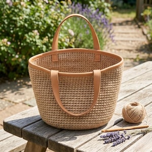 May include: A crocheted tote bag with tan leather handles and trim sits on a wooden table. The bag is a light brown color and has a rounded shape. A ball of twine, crochet hooks, and lavender sprigs are also on the table.