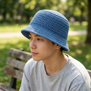 Puede incluir: Un sombrero de pescador de ganchillo azul con un ala ligeramente inclinada hacia abajo. El sombrero está hecho de un material tejido texturizado. La persona de la imagen lleva una camiseta gris.