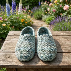 May include: A pair of handmade, blue and green crochet slippers. The slippers are made of a textured yarn and sit on a weathered wooden surface. The background features a blurred garden with colorful flowers.