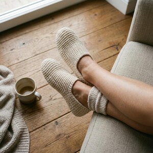 May include: A pair of cream-colored, crocheted slippers are worn by a person relaxing on a beige armchair. The slippers are paired with matching socks. A mug of coffee and a beige blanket rest on the wooden floor nearby, creating a cozy, relaxed atmosphere.