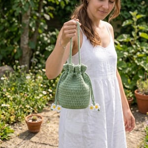May include: A sage green crochet drawstring bucket bag featuring a long braided handle and two decorative white and yellow daisy flower accents attached to the ends of the drawstring cords, creating a textured and functional accessory.