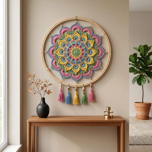 May include: A circular wall hanging featuring a multi-colored crochet mandala mounted on a wooden hoop. The mandala displays concentric layers of pink, yellow, blue, and green yarn. Five hanging tassels in matching colors are attached to the bottom.