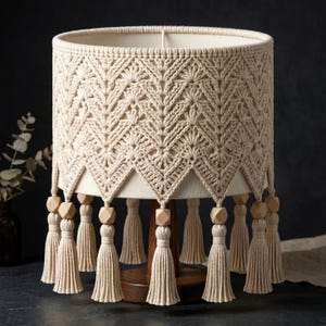 Pode incluir: Abajur de macramé bege com um padrão geométrico e uma franja de borlas. O abajur assenta numa base de madeira escura. As borlas são acentuadas com contas de madeira. O design geral é boémio e decorativo.