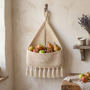 May include: A cream-colored, crocheted fruit basket hanging on a wall, filled with apples, pears, and grapes. The basket has a triangular shape with a fringe along the bottom. The basket is suspended from a hook.