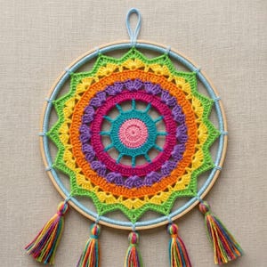 May include: A handmade dreamcatcher with a vibrant, rainbow-coloured crochet design. The circular piece features concentric rings of yellow, orange, pink, purple, and green, with a light blue wooden hoop and tassels. The dreamcatcher is approximately 25 cm in diameter.