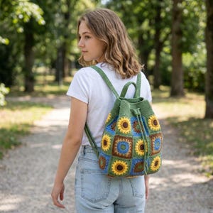 Op de afbeelding: Een groene gehaakte rugzak met een zonnebloemmotief. De tas heeft een trekkoordsluiting en verstelbare bandjes. De zonnebloemvierkanten zijn geel, blauw en bruin. De persoon draagt een wit t-shirt en een blauwe spijkerbroek.