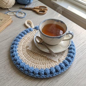 May include: A handmade, round crochet trivet in cream and light blue, with a loop for hanging. A teacup with tea sits on the trivet, along with a saucer and spoon. The trivet is on a light-colored wooden surface, with yarn and scissors in the background.