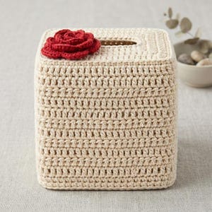 May include: A square, off-white crochet tissue box cover. A red crochet rose is attached to the top. The tissue box cover has a rectangular opening for tissue dispensing. The crochet work has a textured appearance.