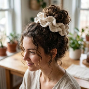 Può includere: Un elastico per capelli all'uncinetto color crema, a balze, indossato in uno chignon. L'elastico è realizzato con un filato spesso e le balze aggiungono volume e consistenza. Lo sfondo comprende una finestra, piante e un tavolo di legno.