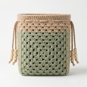 Könnte beinhalten: Eine handgefertigte Häkeltasche in quadratischer Form. Die Tasche hat ein zweifarbiges Design mit einem hellbraunen Oberteil und einem grünen Körper. Die Tasche hat einen Kordelzug mit Holzperlen und ein gewebtes Muster.