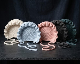 May include: A collection of six crocheted baby bonnets in various pastel colors, including cream, blue, pink, gray, and teal. Each bonnet features a circular design with a ruffled brim and tie strings.
