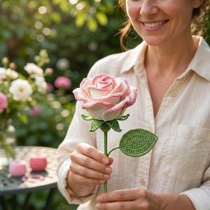 May include: A handmade crochet rose with a white and pink flower head, green stem, and leaf. The rose is held in front of a person wearing a light-colored shirt. The background features a garden setting with other flowers.