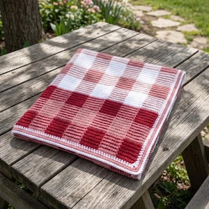 May include: A handmade crochet blanket with a red, white, and pink gingham pattern. The blanket is folded neatly on a wooden picnic table. The blanket's edges are finished with a white border.