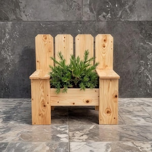 May include: A wooden planter shaped like a small chair, filled with green plants. The planter is made of light-colored wood and has a back with three vertical slats. The chair-shaped planter is on a tiled floor with a dark gray wall in the background.