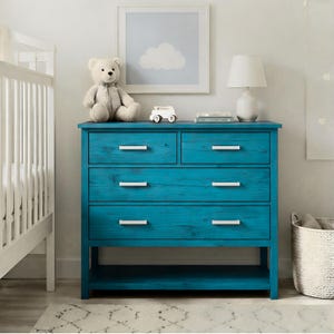 May include: A teal blue wooden dresser with four drawers and white handles. A white teddy bear, toy car, and lamp sit on top. A framed cloud art piece hangs above the dresser, creating a nursery theme.