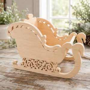 May include: A light-coloured wooden sleigh featuring intricate scrollwork cutouts along the base and curved runners. The structure has a high backrest and open sides with decorative scalloped edges, showcasing a natural wood grain finish throughout.