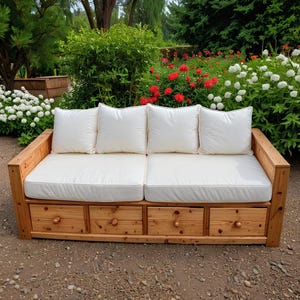 Può includere: Un divano da esterno in legno con quattro cuscini bianchi e quattro cassetti. Il divano è realizzato in legno chiaro e ha un design semplice e rustico. I cuscini sono quadrati e i cassetti hanno pomelli rotondi in legno. Il divano è in un giardino.