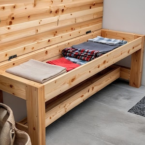 Könnte beinhalten: Eine Holz-Aufbewahrungsbank mit klappbarem Rücken und einem unteren Regal. Die Bank ist aus hellem Holz gefertigt und enthält gefaltete Decken und Handtücher in verschiedenen Farben, darunter Rot, Grau und Blau. Die Bank steht auf einer grauen Betonoberfläche.