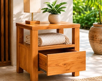 May include: A light brown wooden nightstand with a square top, a lower shelf, and one open drawer. The nightstand features a rectangular lamp with a beige shade and a green potted plant on top, with a folded white textured blanket on the shelf.