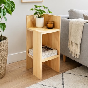 May include: A light-colored wooden corner side table with two shelves. The top shelf holds a potted plant and a candle. The bottom shelf displays books. The table is next to a gray sofa and a woven basket with a plant.