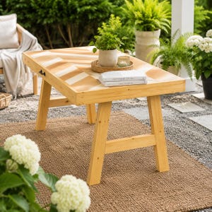 May include: A rectangular wooden coffee table with a light-colored chevron pattern on the tabletop. The table features four angled legs and a small black metal handle on the side. Two stacked books titled Outdoor Living and This Is Home sit on top.