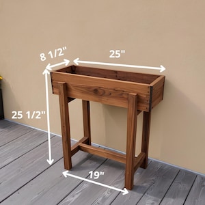 May include: A wooden raised garden planter with a rectangular trough, 25 inches long, 8.5 inches wide, and 25.5 inches tall. The planter is supported by four legs, 19 inches wide. The wood has a natural finish.