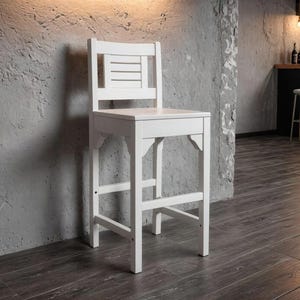 May include: A tall, white wooden bar stool with a slatted backrest and a square seat. The stool has four legs and a footrest. The stool is painted white and stands on a dark wood floor.