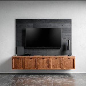 May include: A modern entertainment center featuring a large flat-screen TV mounted on a dark wood panel. Below, a floating wooden cabinet with six doors provides storage. A black speaker and lamp complete the setup. The cabinet is approximately 72 inches wide.