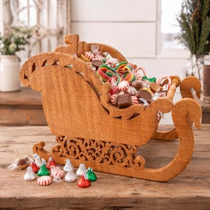 May include: A light brown wooden sleigh with intricate scrollwork along the base, filled with a variety of colorful candies including rainbow gummy strips, peppermint rounds, and chocolate pieces, with more candies scattered around the base.