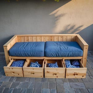 Könnte beinhalten: Outdoor-Sofa aus Holz mit zwei blauen Kissen und vier Ausziehschubladen. Die Schubladen sind mit gemusterten Kissen gefüllt. Das Sofa ist aus hellem Holz gefertigt und für den Außenbereich konzipiert.